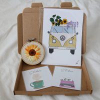 overview of a sample mystery box containing a 3inch sunflower embroidery hoop, camper van print, tea sticker and typewriter sticker