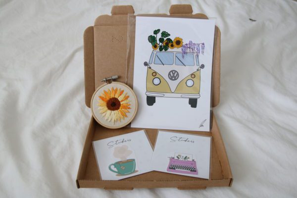 overview of a sample mystery box containing a 3inch sunflower embroidery hoop, camper van print, tea sticker and typewriter sticker
