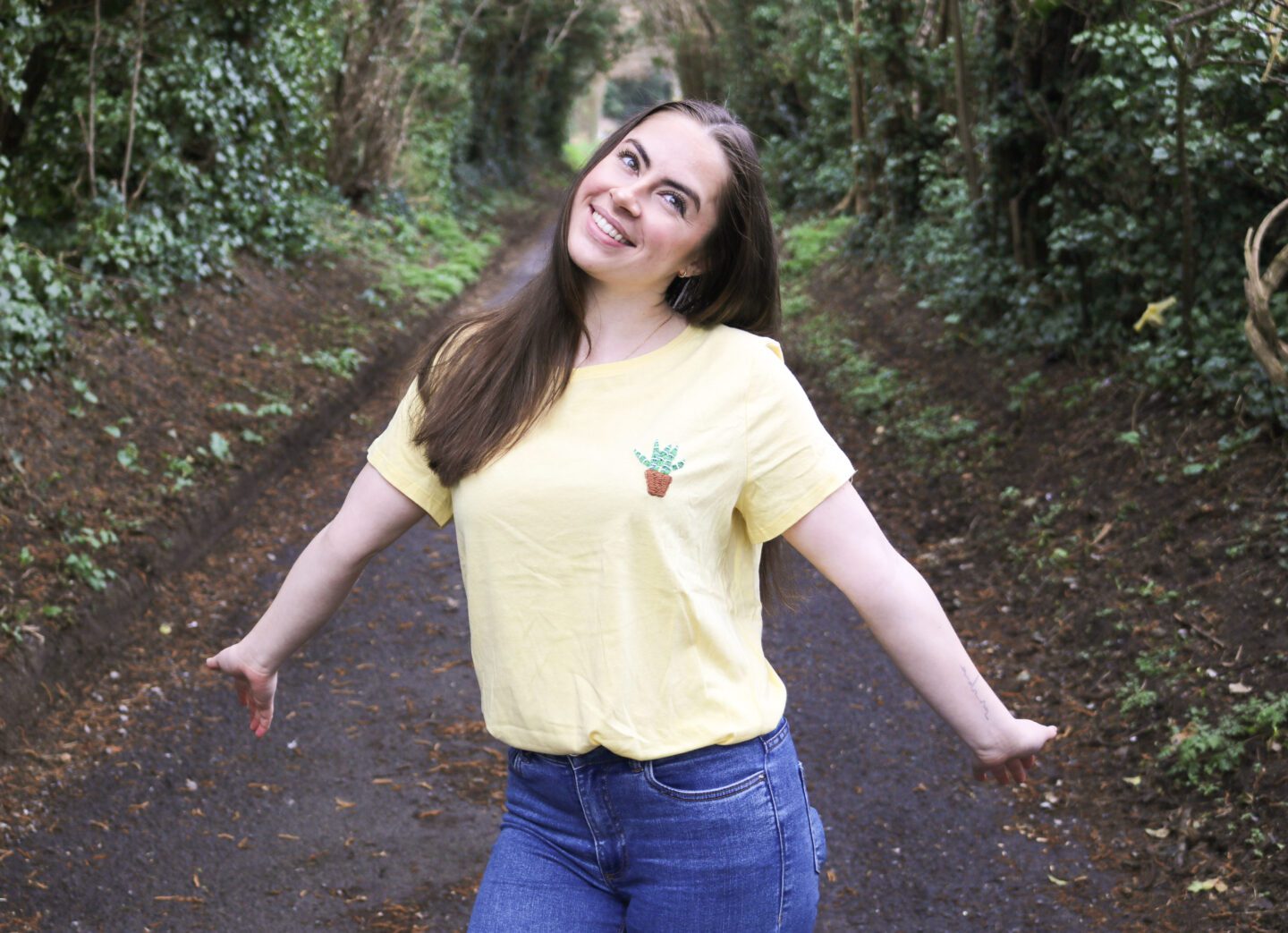 A brunette girl wearing blue skinny jeans and a yellow t-shirt hand embroidered with an Aloe vera plant