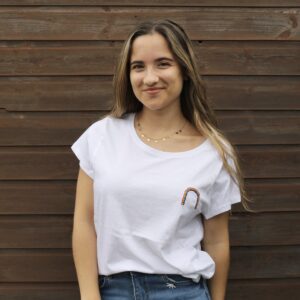 A brunette girl stood in front of a brown fence wearing blue jeans and a white top with a hand embroidered rainbow