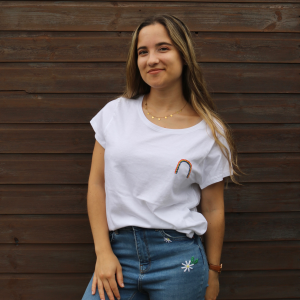 A brunette girl wearing. white t-shirt hand embroidered with a rainbow paired with blue denim jeans hand embroidered with daisies