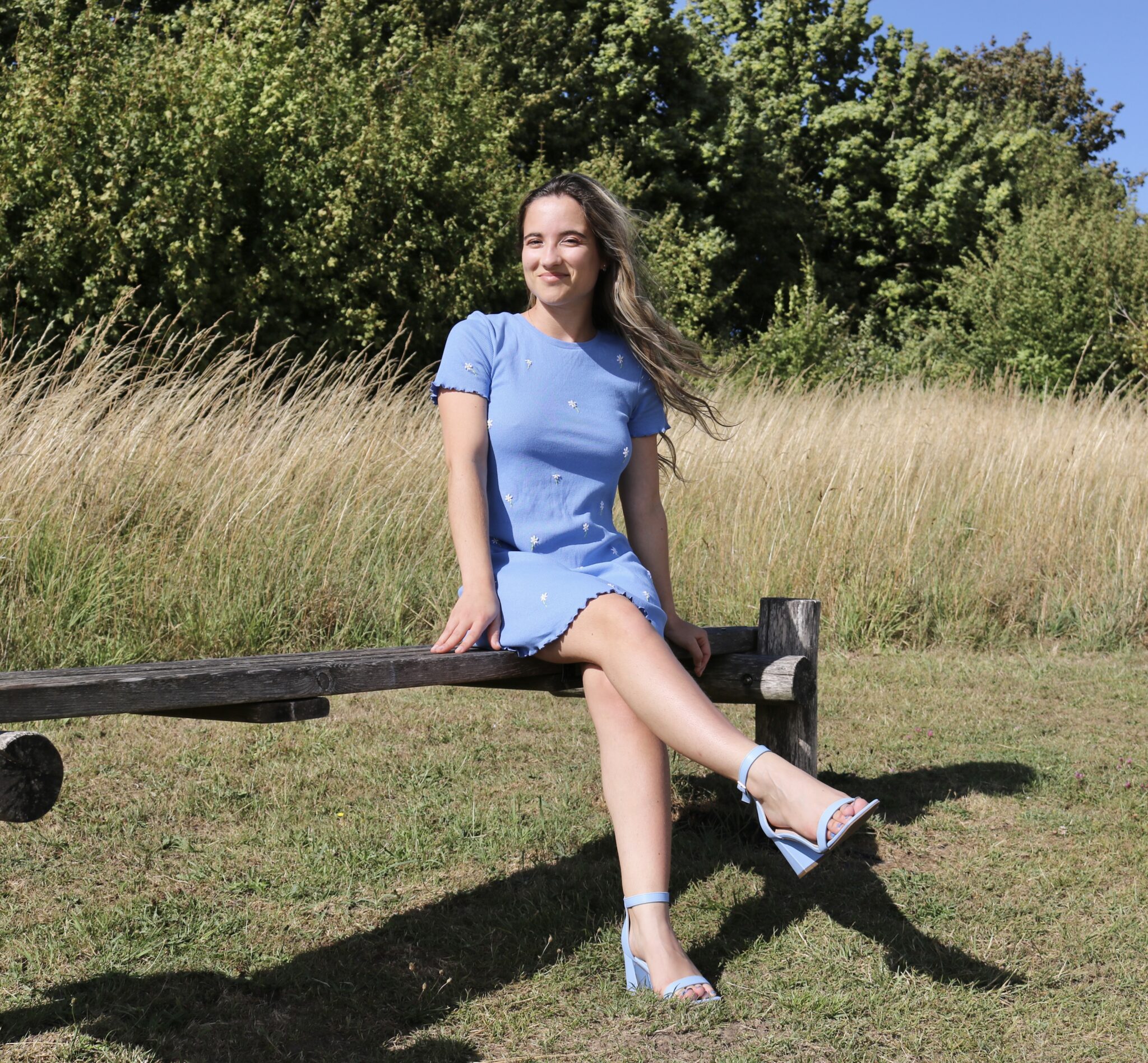 A brunette girl sat on a park bench wearing a blue body con dress hand embroidered with daisies