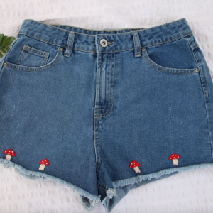 A girl wearing blue denim shorts hand embroidered with 4 mushrooms across the hem of the shorts