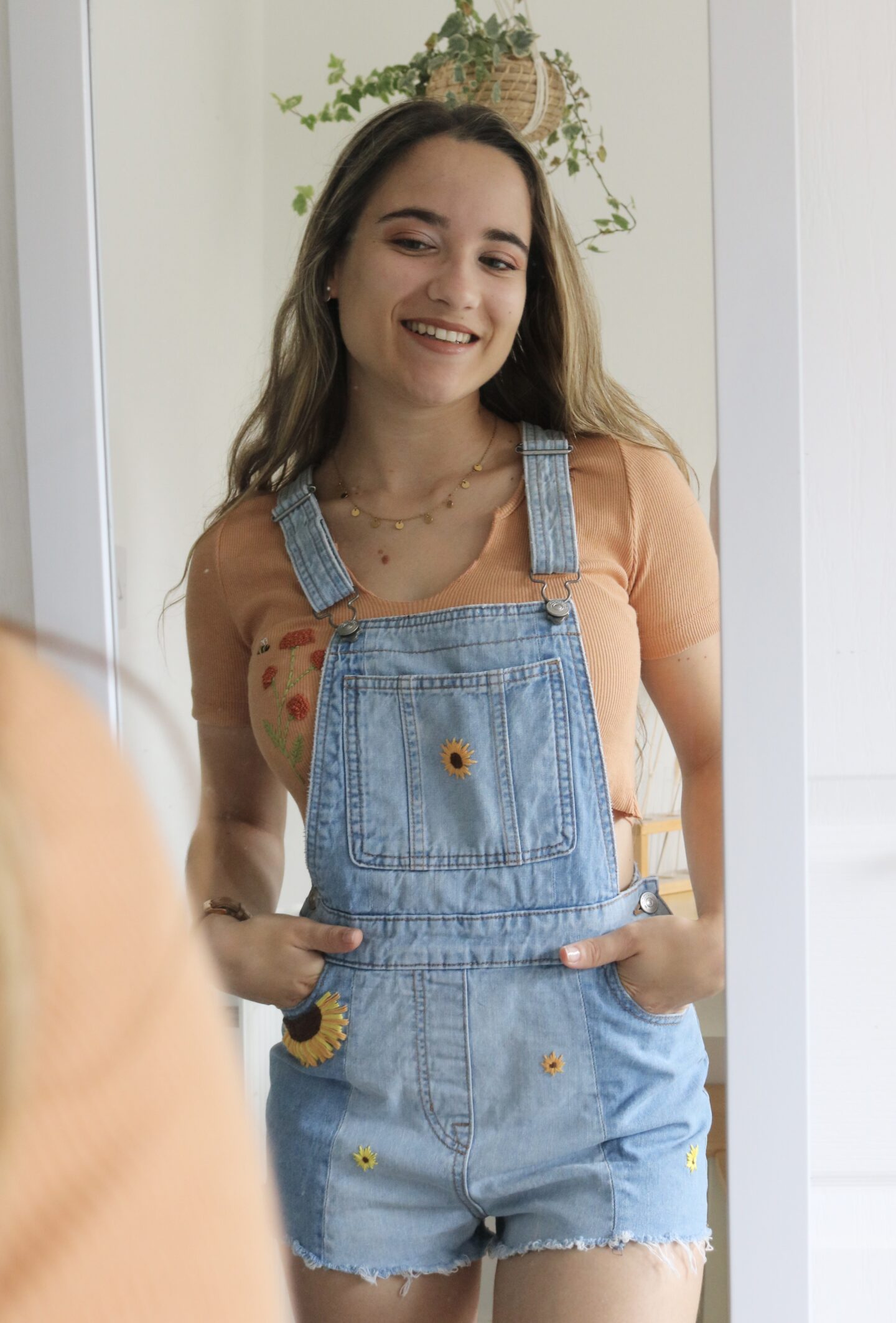 A brunette girl looking in the mirror wearing blue overall shorts hand embroidered with sunflowers