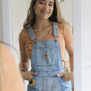 A brunette girl looking in the mirror wearing blue overall shorts hand embroidered with sunflowers