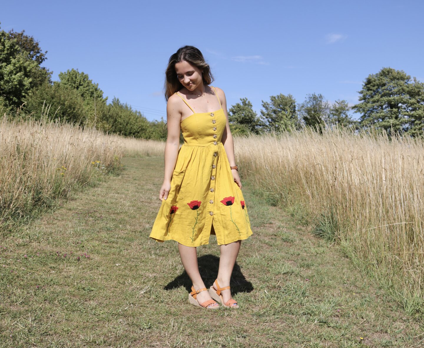 A brunette girl stood in a field wearing a yellow knee length button up dress hand embroidered with 4 red poppies at the bottom of the skirt