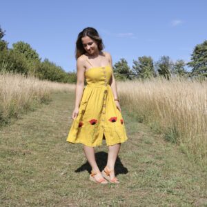 A brunette girl stood in a field wearing a yellow knee length button up dress hand embroidered with 4 red poppies at the bottom of the skirt