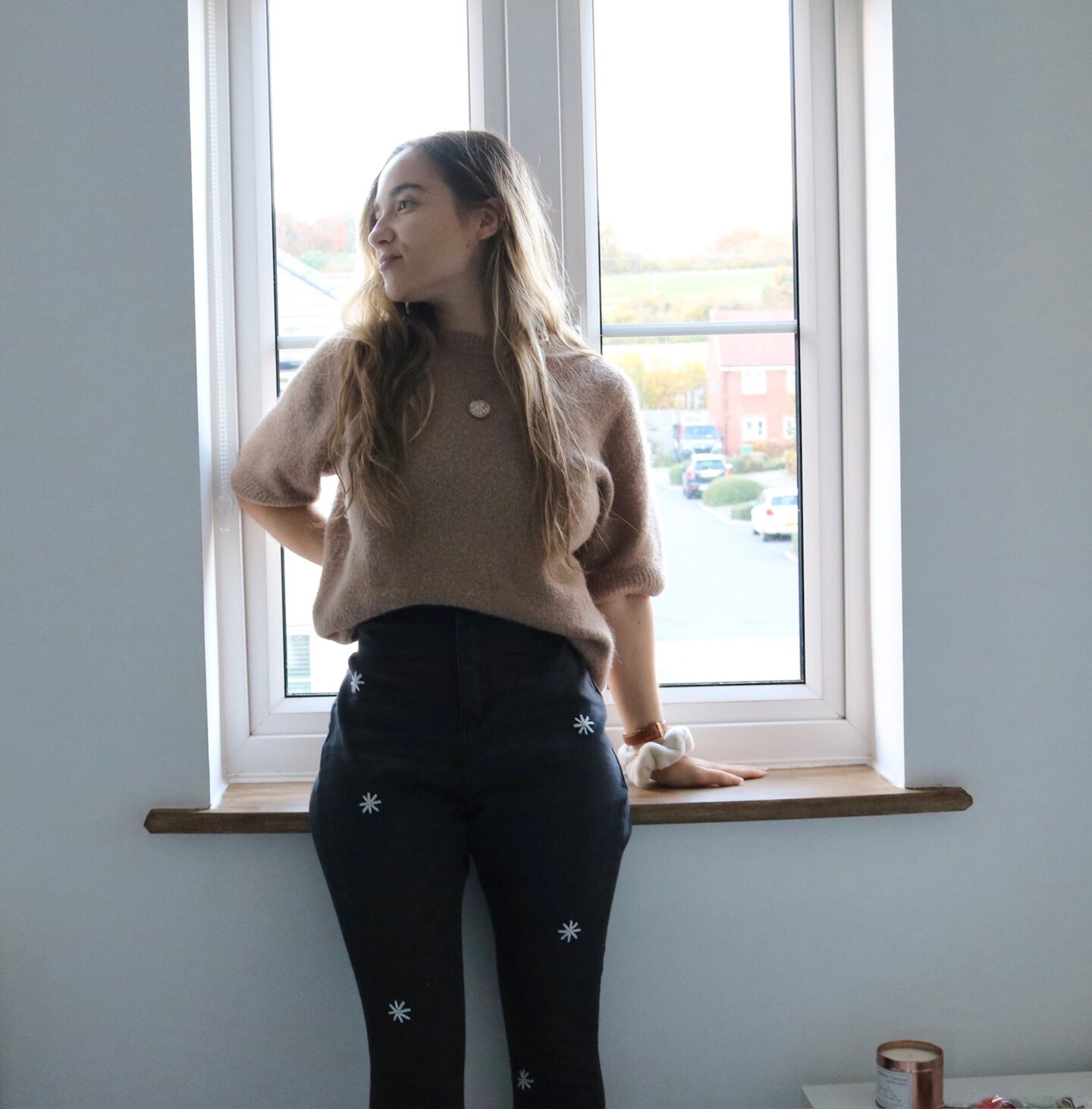 A brunette girl standing by a window wearing a brown jumper and black skinny jeans hand embroidered with scattered white daisies