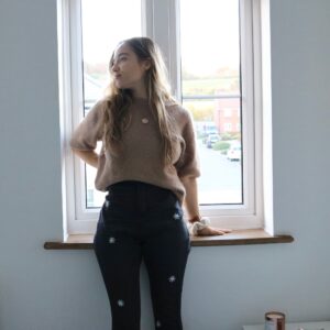 A brunette girl standing by a window wearing a brown jumper and black skinny jeans hand embroidered with scattered white daisies