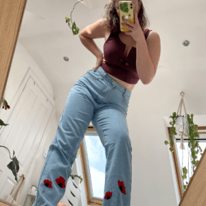 brunette girl taking a mirror selfie wearing light blue mom jeans hand embroidered with 4 big poppies growing up the hem