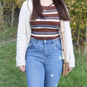 A lifestyle shot of a woman wearing blue skinny jeans hand embroidered with white daisies down one leg paired with a brown stippy t-shirt and white cardigan
