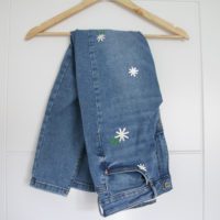 Jeans folded over a hanger. The jeans are blue with white hand embroidered daisies on them