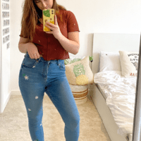 brunette model taking a mirror selfie standing up wearing blue skinny jeans with embroidered daisies down one leg