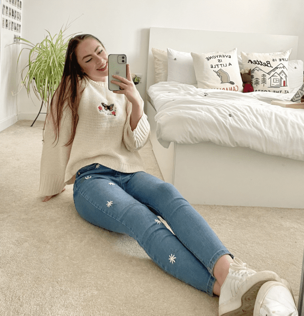 brunette girl taking a mirror selfie wearing blue skinny jeans and embroidered with white daisies down one leg