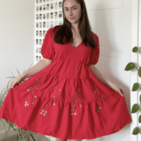 A brunette girl wearing a red floaty dress hand embroidered with blue flowers on vines.