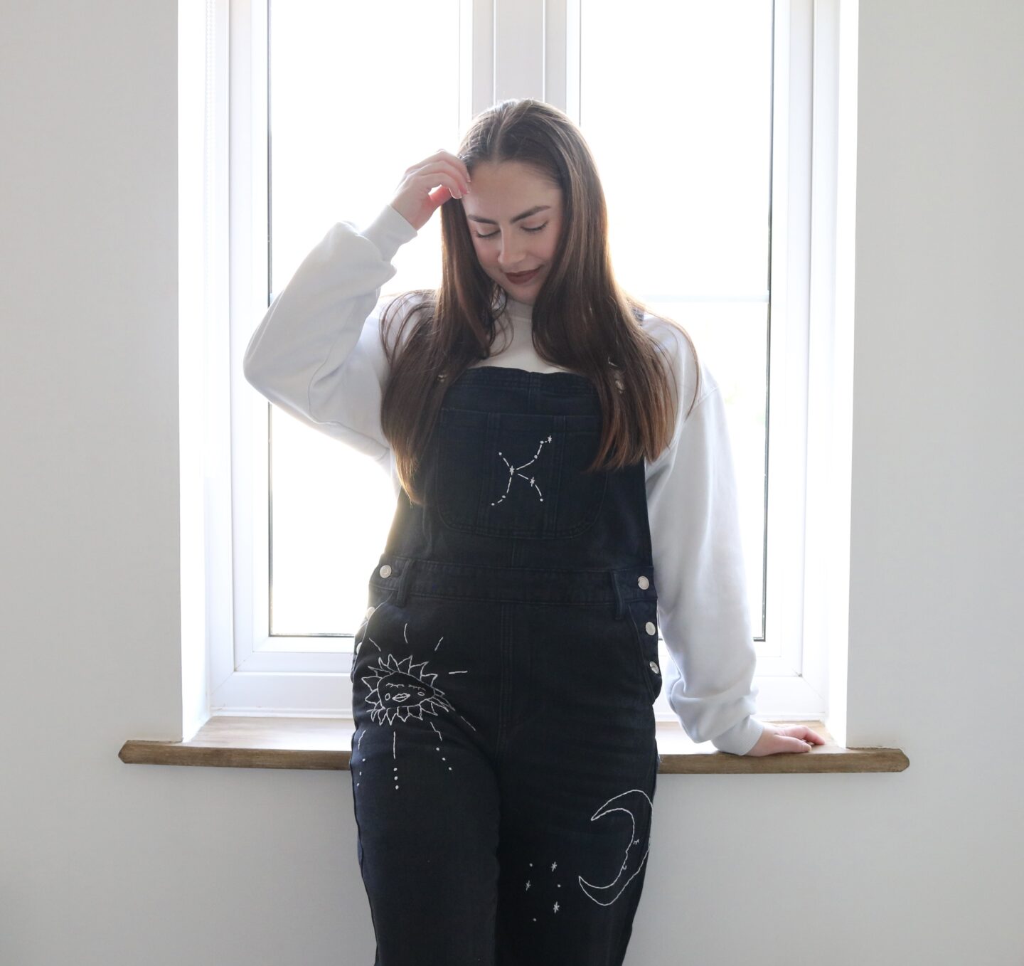 A brunette woman stood by a window wearing black overalls hand embroidered with a white outline of a constellation, moon, sun, and stars