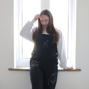 A brunette woman stood by a window wearing black overalls hand embroidered with a white outline of a constellation, moon, sun, and stars