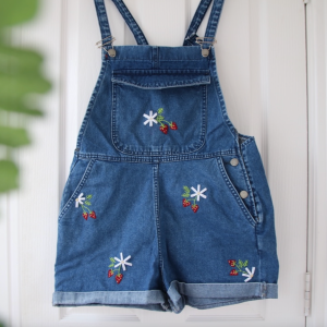 A blue pair of short overalls hand embroidered with strawberries and daisies