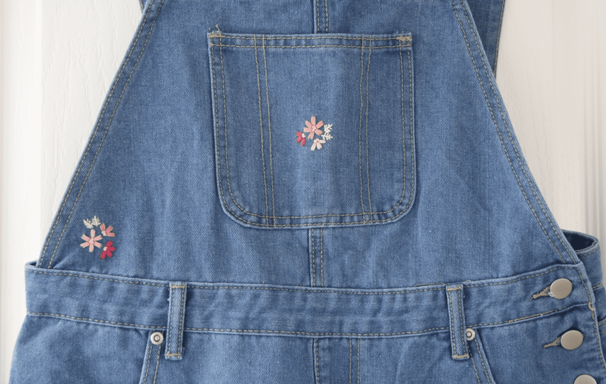 The front pocket of a pair of denim short overalls hand embroidered with pink blossom