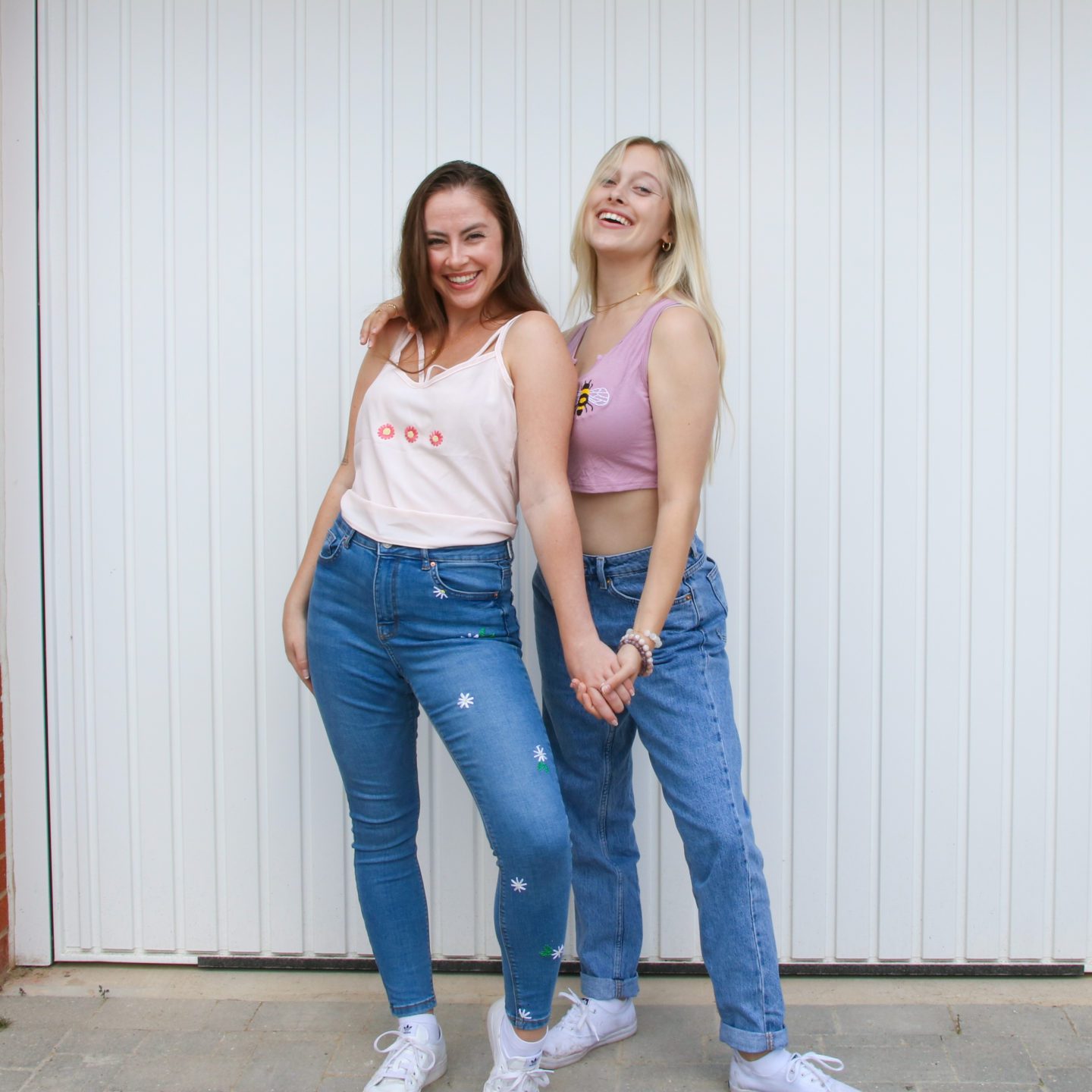 two females wearing hand embroidered clothing. Outfits include: a pink top embroidered with daisies, blue jeans embroidered with daisies, and a pink top embroidered with a bee paired with plain blue jeans