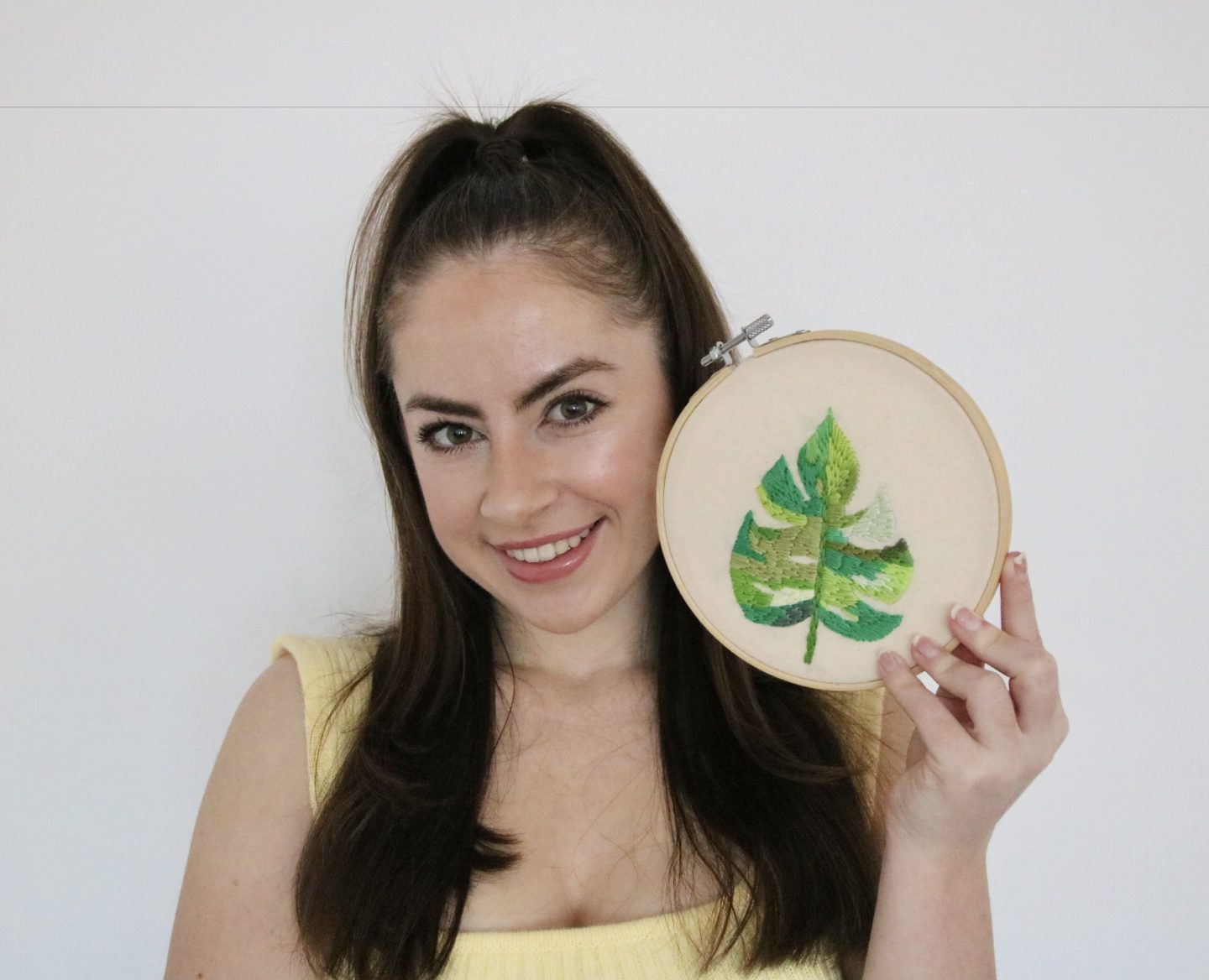 A brunette girl smiling and holding a 6 inch embroidery hoop hand embroidered with a green monstera leaf
