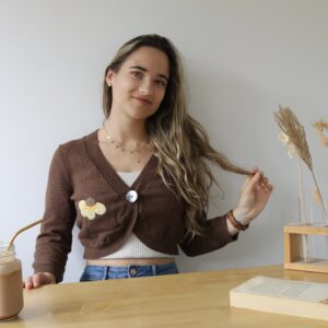 A brunette girl sat at a table reading a book with an iced coffee wearing a brown cardigan hand embroidered with a yellow flower