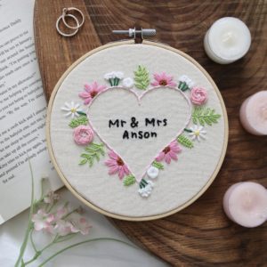 6 inch hoop hand embroidered with pink and white flowers in the shape of a heart. In the middle is custom text reading "Mr & Mrs Anson"