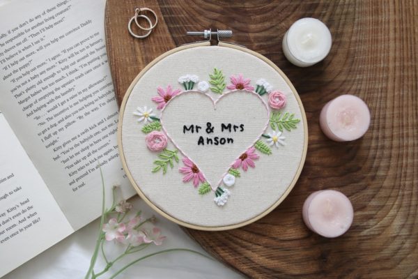 6 inch hoop hand embroidered with pink and white flowers in the shape of a heart. In the middle is custom text reading "Mr & Mrs Anson"