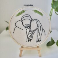 A 6 inch embroidery hoop on a wooden stand hand embroidered with the outline of a boyfriend and girlfriend