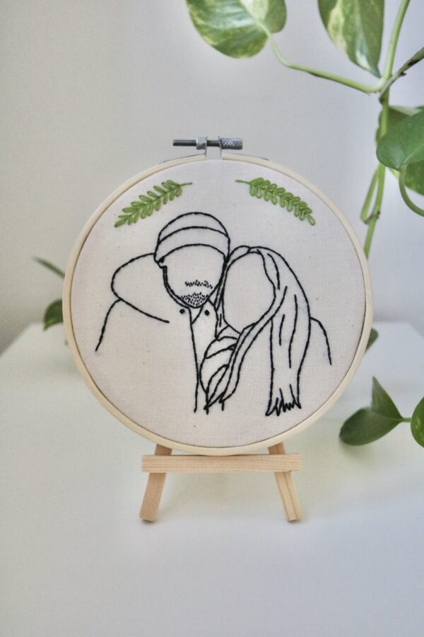 A 6 inch embroidery hoop on a wooden stand hand embroidered with the outline of a boyfriend and girlfriend