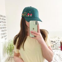 brunette model wearing the forest green cap with hand embroidered mushrooms, bees, and green vines with orange roses