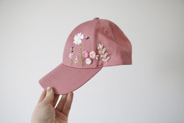 pink cap hand embroidered with a big white flower, daisy, pink roses, green leaves and vines, and bees