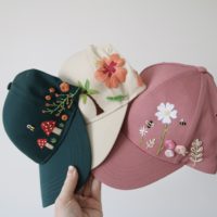 3 caps, a green one embroidered with toadstools, cream hat embroidered with palm trees and hibiscus flowers and pink hat embroidered with flowers and bees
