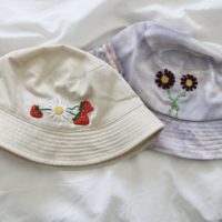 Overview of two bucket hats. First hat is cream with a hand embroidered daisy and strawberries. Second hat is purple with cosmos flowers