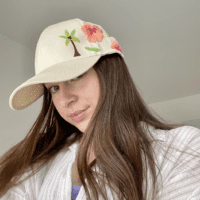 Brunette model wearing a cream cap hand embroidered with a palm tree and pink hibiscus flower