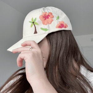 brunette model wearing a cap, side view showing a cream cap hand embroidered with a palm tree and pink hibiscus flowers