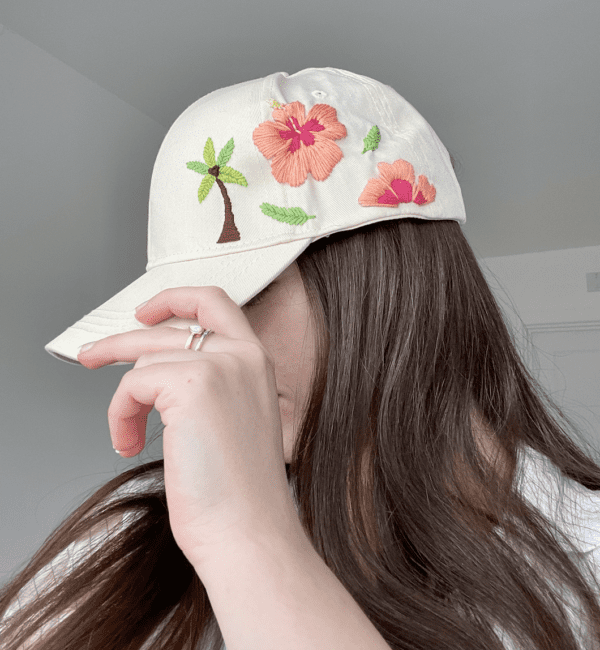 brunette model wearing a cap, side view showing a cream cap hand embroidered with a palm tree and pink hibiscus flowers