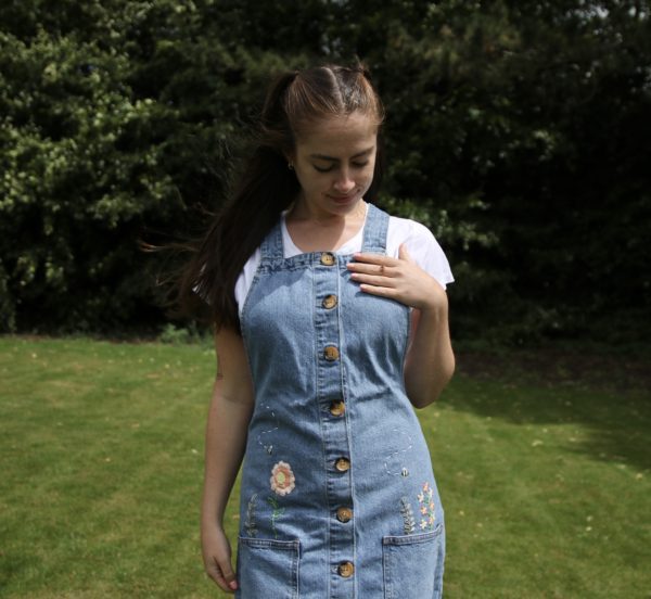 close up of the girl wearing a blue denim pinafore dress hand embroidered with pink flowers, greenery, and bees