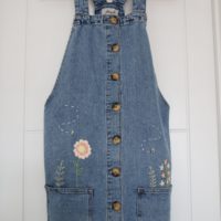 blue denim pinafore dress hand embroidered with pink flowers, and greenery growing from the pockets, and 2 bees flying around
