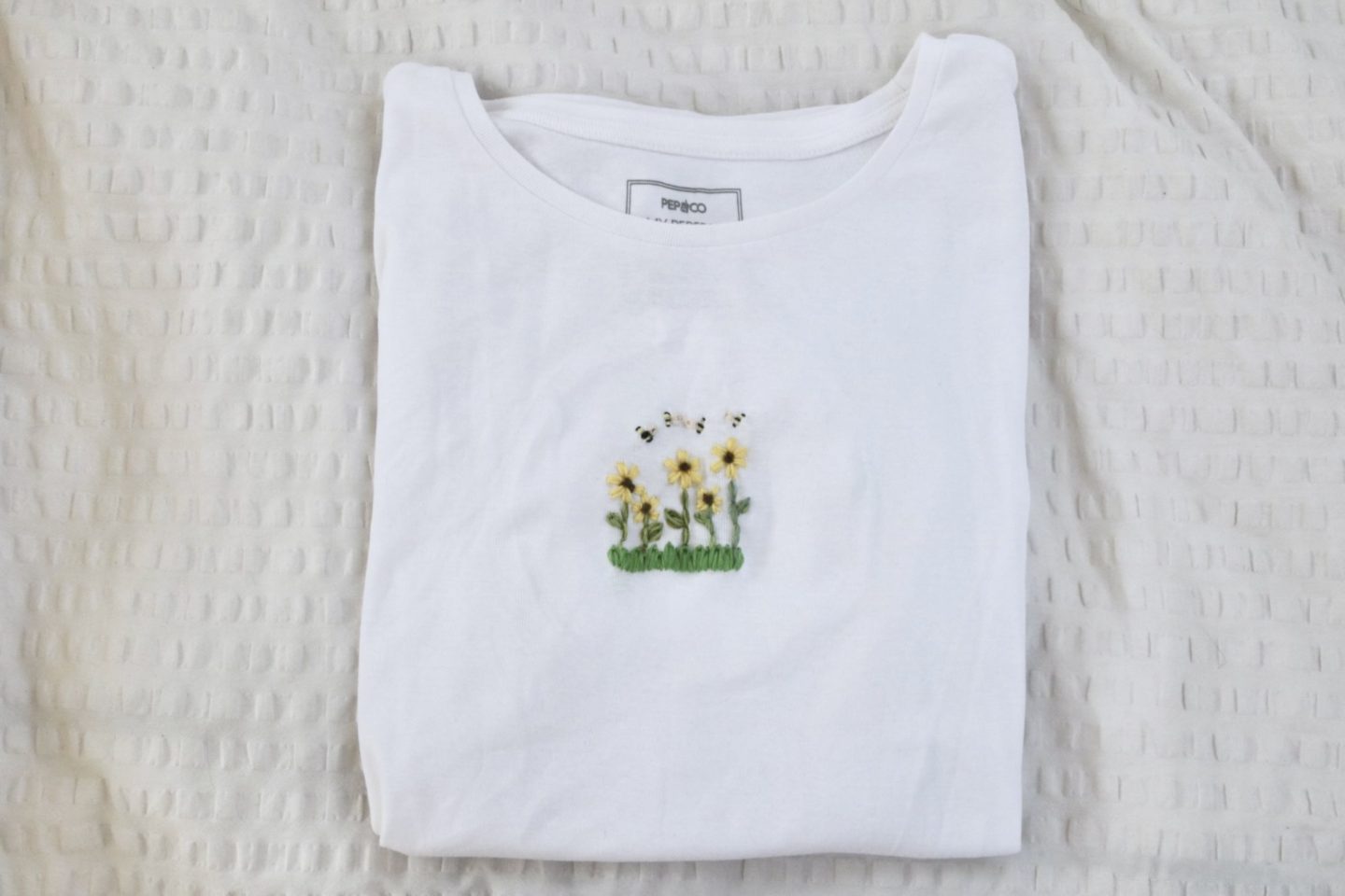 A white t-shirt hand embroidered with grass, sunflowers, and bees in the centre of the top