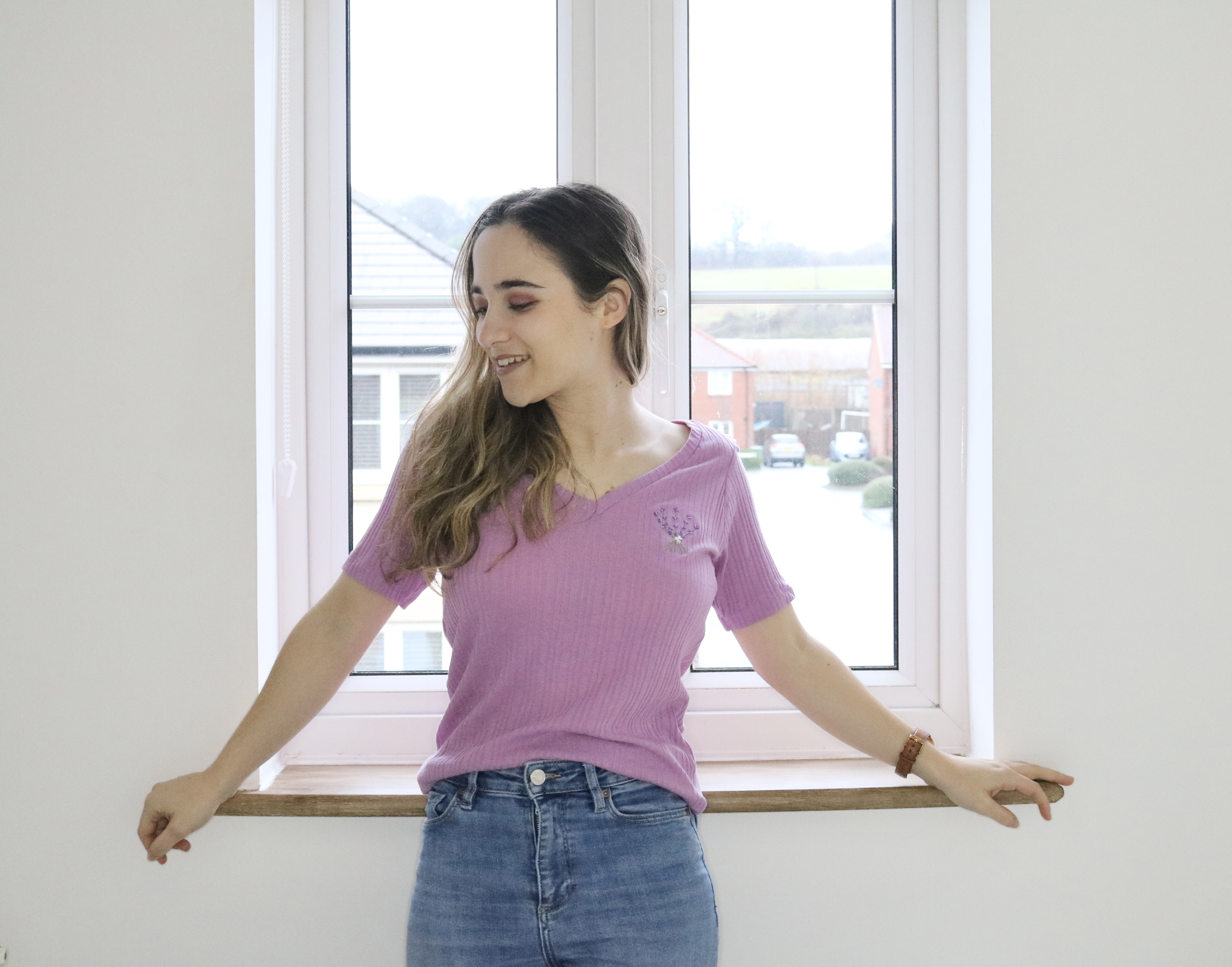 A brunette girl stood by a window wearing a purple top hand embroidered with lavender