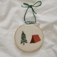 A 3 inch embroidery hoop hand embroidered with an orange tent and tree