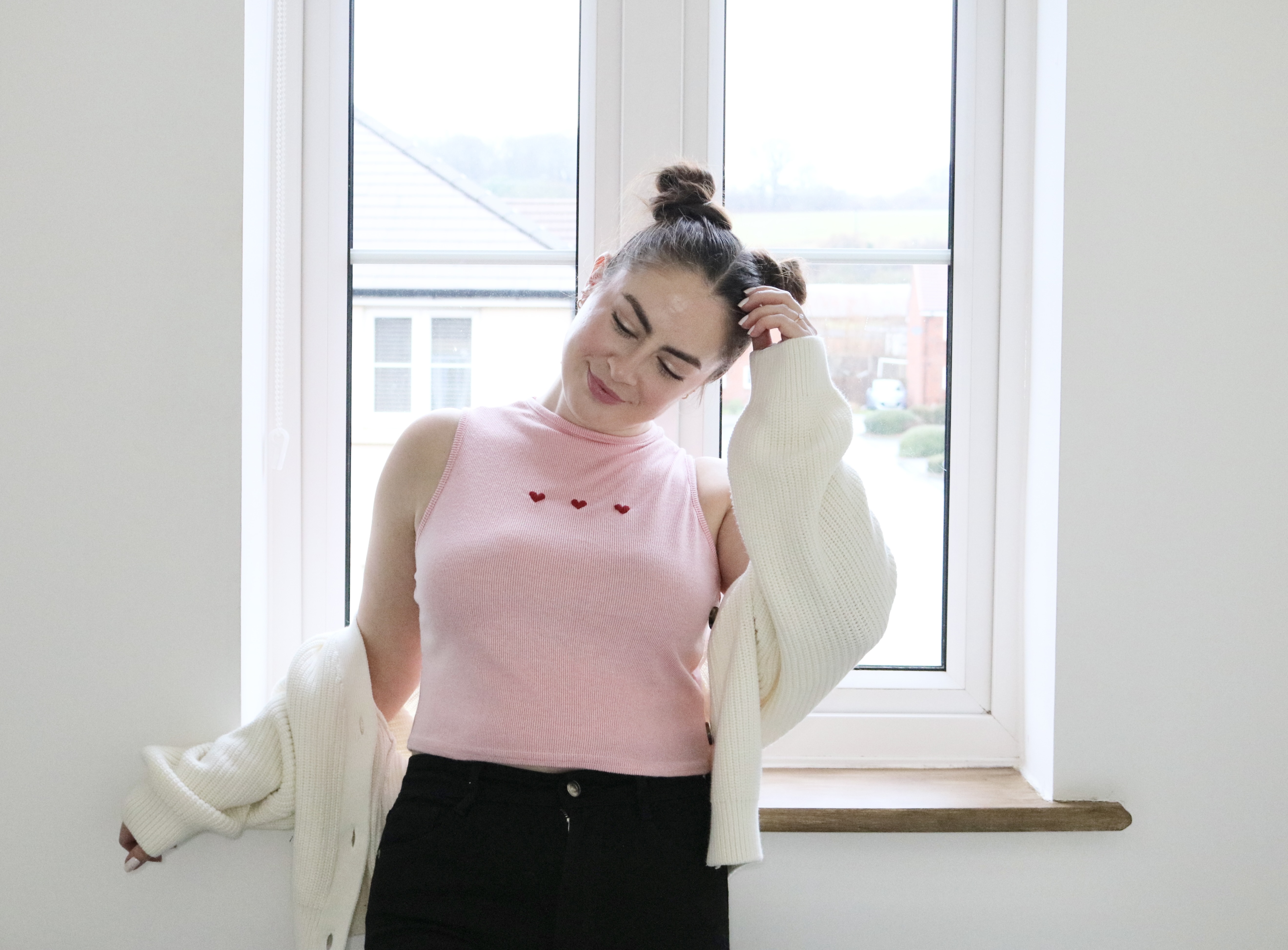 A brunette girl standing by a window wearing black jeans, a white cardigan, and a pink top hand embroidered with three red hearts