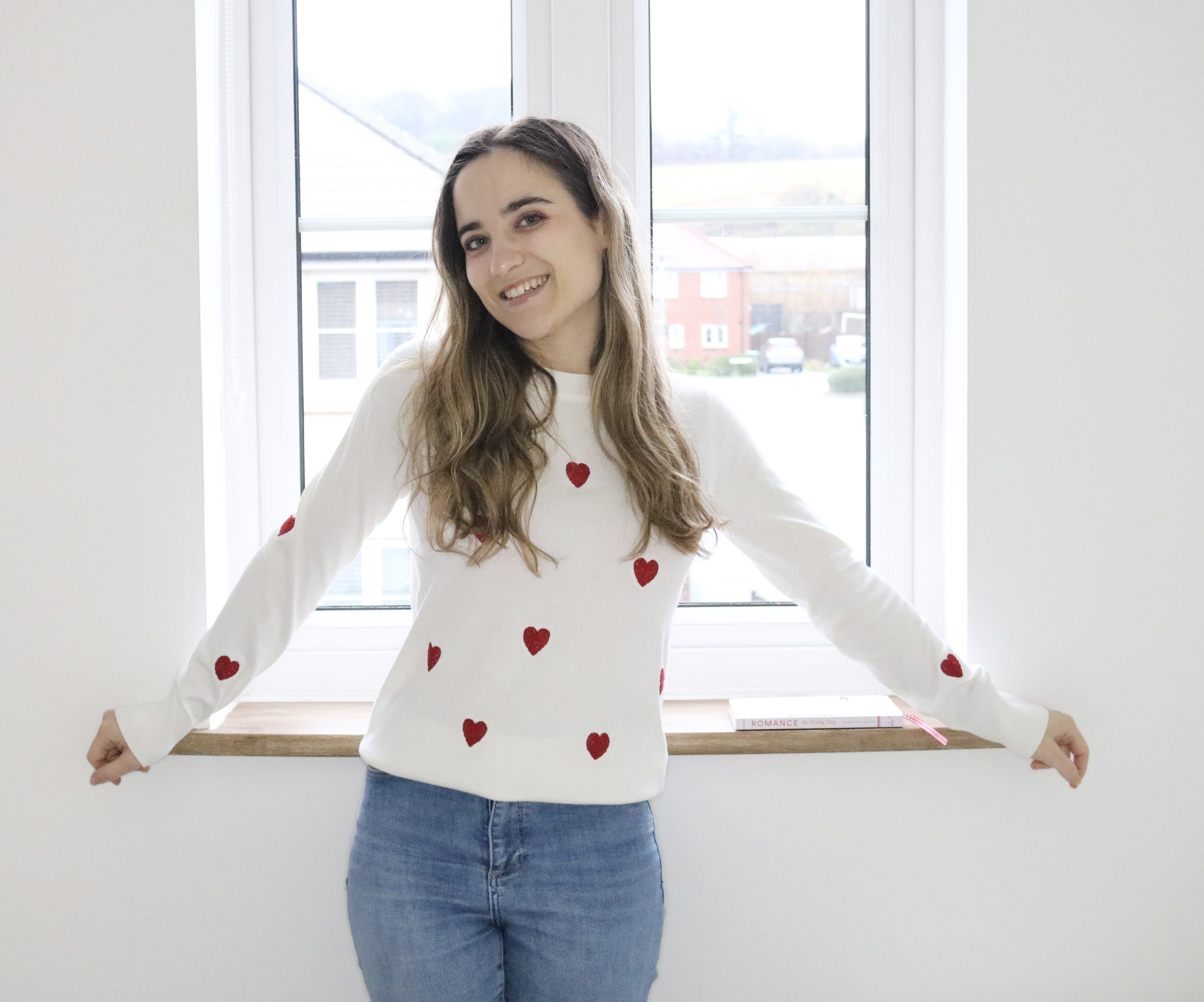A brunette girl stood by a window wearing blue jeans and a white jumper with hand embroidered red hearts