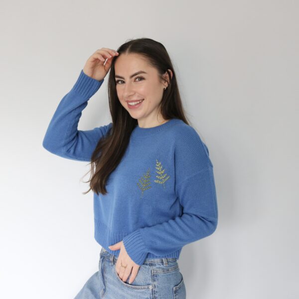 Brunette girl smiling and playing with her hair wearing an electric blue jumper hand embroidered with two fern leaves