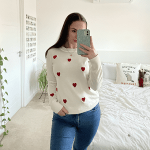 A brunette girl taking a mirror selfie wearing a white jumper hand embroidered with red hearts and blue jeans