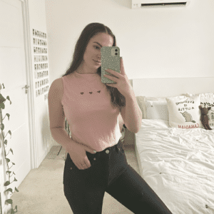 A brunette girl taking a mirror selfie wearing a pink and white top hand embroidered with three red hearts in the centre