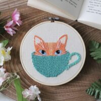 A hand embroidered cat poking its head out of a cup