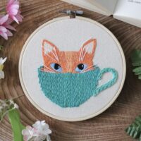 a 6 inch hoop hand embroidered with a kitten face poking out of a mug