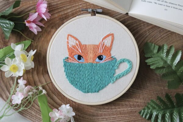 a 6 inch hoop hand embroidered with a kitten face poking out of a mug